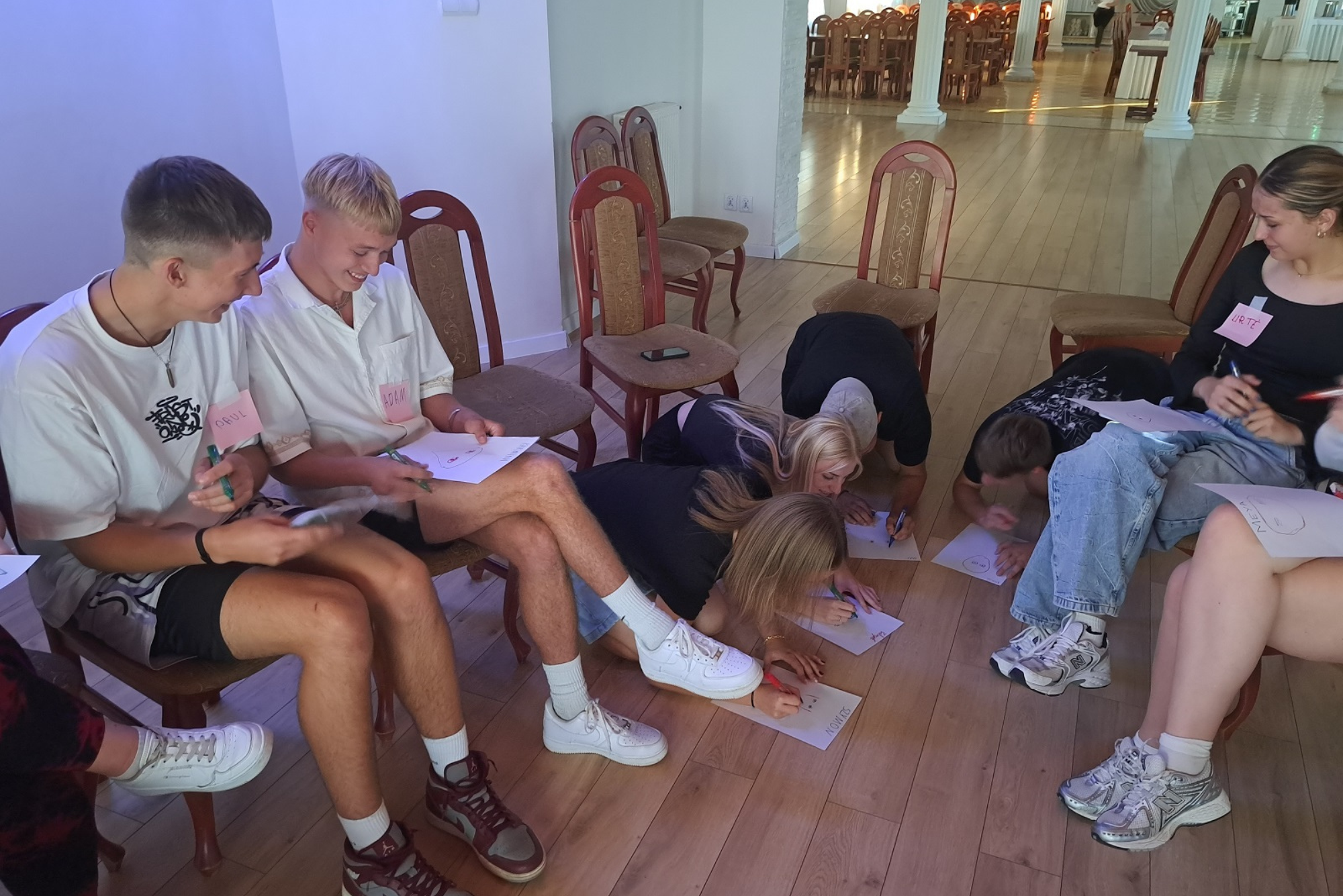 Młodzież siedzi na krzesłach lub na podłodze i rysuje coś na kartkach papieru