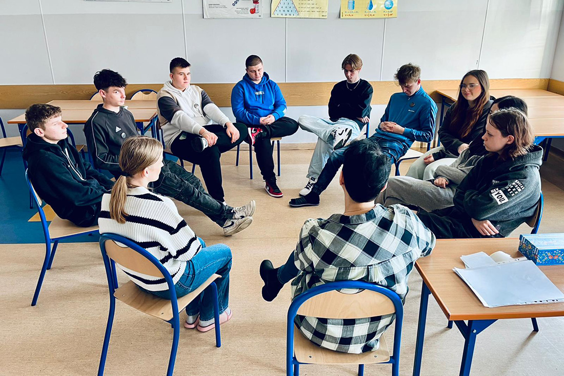 Na zdjęciu widać grupę młodzieży w trakcie zajęć warsztatowych siedzącą na krzesłach w okręgu.