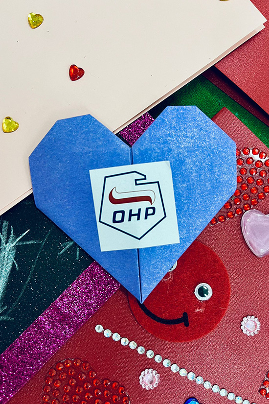 Na zdjęciu widać niebieskie serduszko origami z logiem OHP na tle kartek z życzeniami.