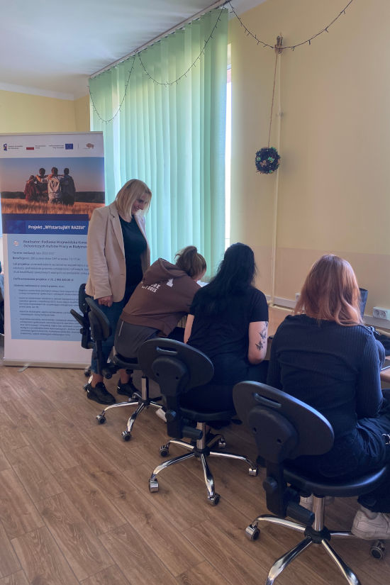 Zdjęcie przedstawia młodzież siedzącą przy biurkach. W tle widać rollup promujący projekt "WYstartujMY RAZEM"